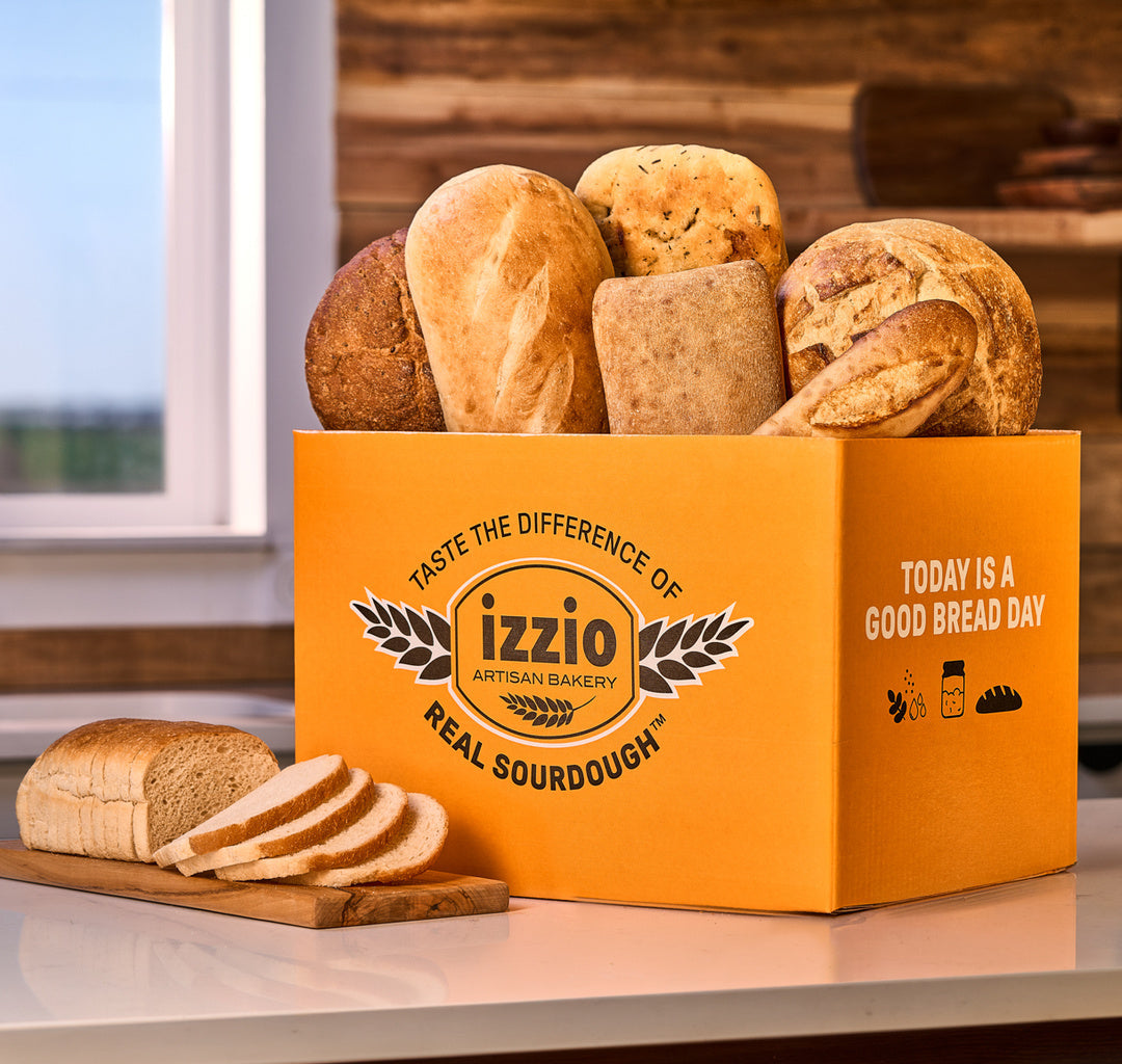Link to The Real Sourdough Edit by izzio. An orange Izzio Artisan Bakery cardboard box filled with assorted artisan sourdough bread loaves and slices, displayed on a kitchen counter with a loaf and slices in front.
