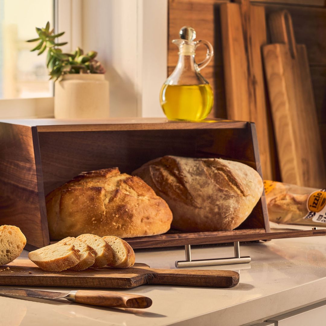 A dark wood bread box with metal handles stands on a kitchen counter, filled with rustic loaves of bread. Sliced bread, a glass bottle of olive oil, and a small potted plant sit nearby in a warm, sunlit kitchen setting. Links to The Real Sourdough Edit