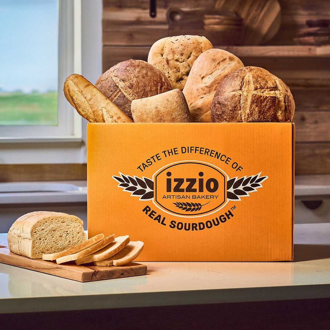 An orange cardboard box labeled 'izzio Artisan Bakery Real Sourdough' filled with assorted rustic sourdough loaves, placed on a kitchen counter with sliced bread and a loaf in front. A window and wooden shelves are visible in the background.