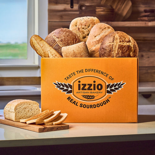An orange cardboard box labeled 'izzio Artisan Bakery Real Sourdough' filled with assorted rustic sourdough loaves, placed on a kitchen counter with sliced bread and a loaf in front. A window and wooden shelves are visible in the background.