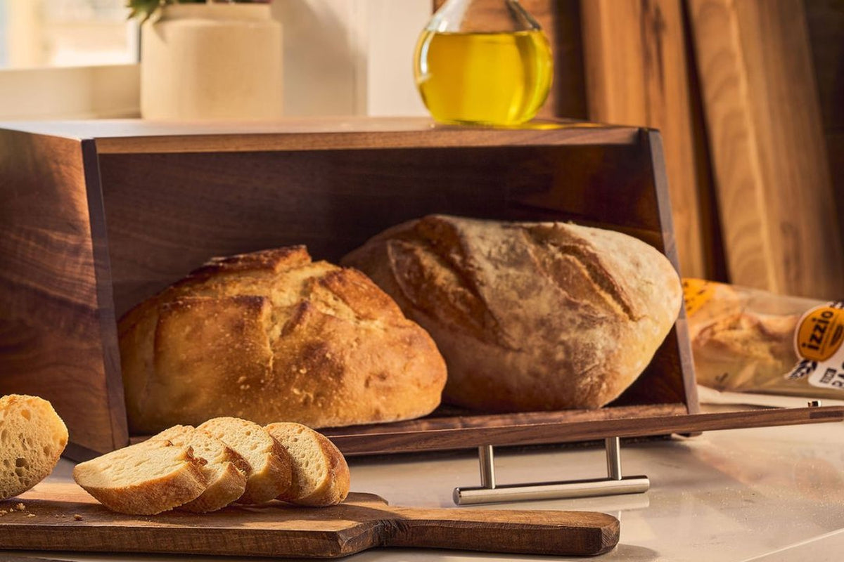 A wooden bread box with a sliding pull-out tray holding two rustic loaves of bread, with sliced bread and a bottle of olive oil in a warm kitchen setting.