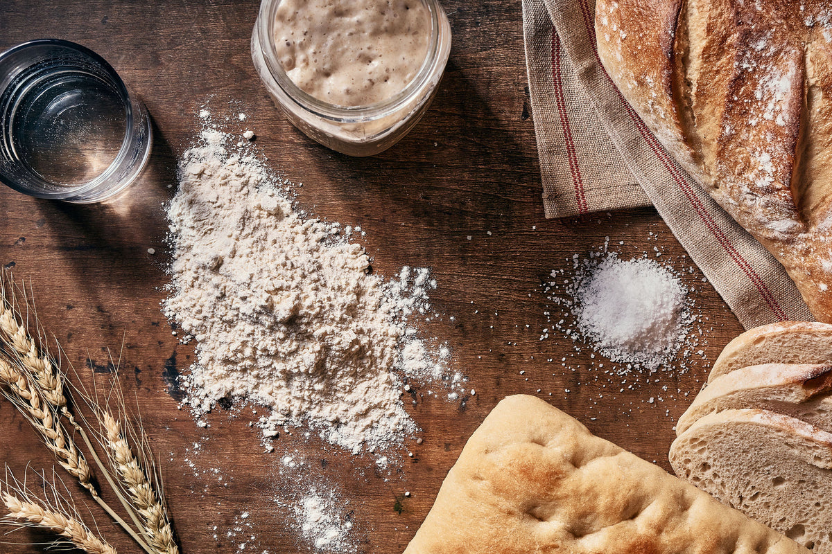 A styled flat-lay of bread and baking ingredients on a dark wooden table, including sliced bread, a loaf, flour, salt, wheat stalks, and a glass of water.